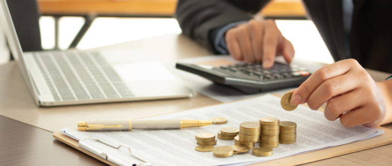 A imagem mostra uma pessoa cuidando das finanças, representando a gestão financeira de uma transportadora. Ela está sentada e sobre a mesa tem um notebook, uma calculadora e uma prancheta com uma planilha. Sobre essa planilha tem uma caneta e várias pilhas de moedas. A pessoa está fazendo contas na calculadora e contando as moedas.
