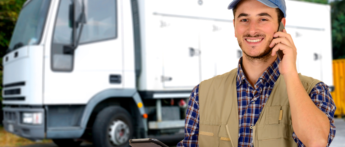 Caminhoneiro falando ao celular, com tablet na mão e caminhão branco ao fundo, ilustra o cadastro e a consulta de motoristas.