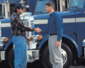 Dois homens apertam as mãos em frente a uma frota de caminhões, mostrando como negociar fretes do jeito certo.