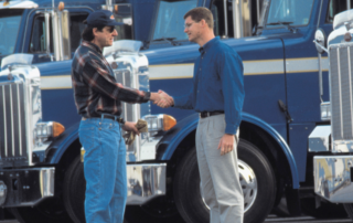 Dois homens apertam as mãos em frente a uma frota de caminhões, mostrando como negociar fretes do jeito certo.