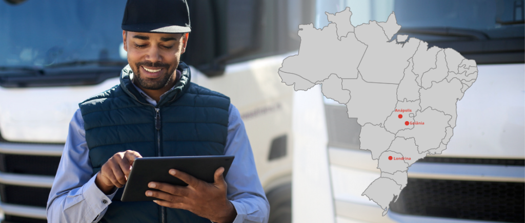 Motorista de caminhão usando tablet em garagem, com mapa do Brasil ao lado, destacando as cidades de Anápolis, Goiânia e Londrina.