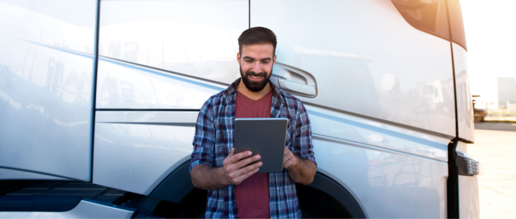 Um homem está conferindo informações num tablet. Ele está apoiado em seu caminhão ou carreta, que está estacionado num pátio.