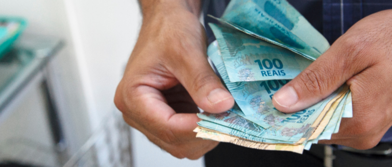 Homem contando notas de dinheiro. Ele segura várias notas em suas mãos. A maior parte é de notas de 100 reais e tem algumas cédulas de 20 reais.