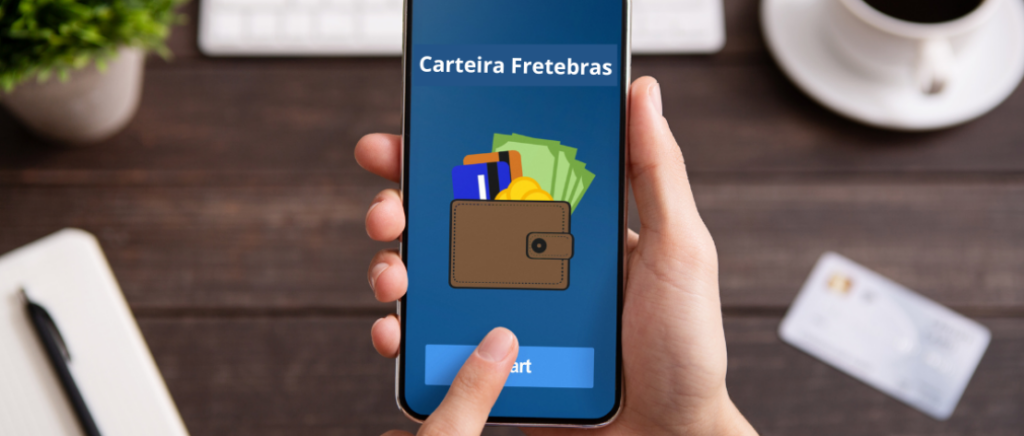 Uma pessoa segura um celular. Ela vai acessar a Carteira Fretebras, que já aparece na tela. Abaixo, aparece uma mesa com um vaso de planta, computador, xícara com café, caderno, caneta e um cartão.