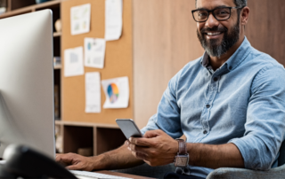 Homem está em um escritório. Ao fundo tem uma estante e um painel com vários gráficos. Ele está sentado, em frente ao seu computador. Sua mão direita está sobre o mouse e a outra está segurando o celular.