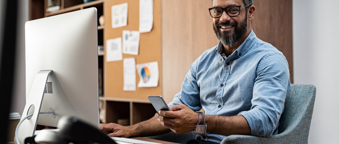 Homem está em um escritório. Ao fundo tem uma estante e um painel com vários gráficos. Ele está sentado, em frente ao seu computador. Sua mão direita está sobre o mouse e a outra está segurando o celular.