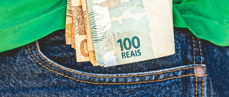 Um homem está vestindo uma calça jeans e uma camiseta verde. Ele está colocando no bolso algumas notas de dinheiro, representando a economia do caminhoneiro na rodagem.