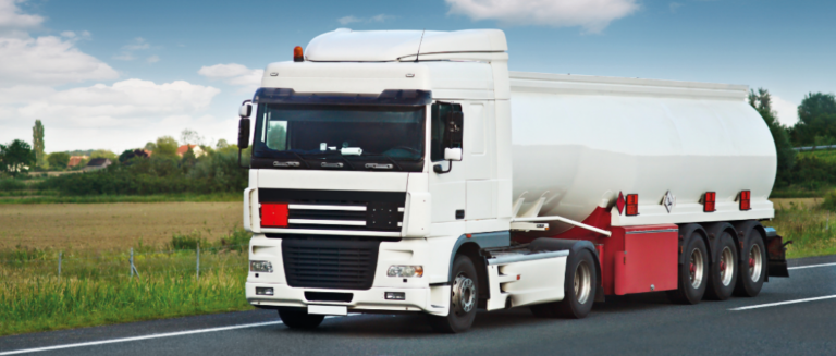 Um caminhão branco com uma carroceria silo está trafegando em uma rodovia. A pista está livre, sem nenhum outro veículo. Ao fundo, tem uma paisagem com grama e árvores.