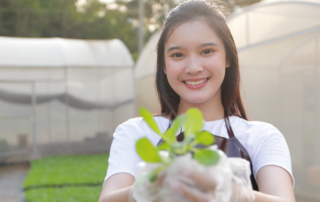 Uma mulher está sorrindo e segurando a muda de uma planta que está crescendo. Ela veste uma camiseta branca e um avental marrom. Ao fundo, tem duas estufas, representando o cultivo de alguns produtos do agronegócio no Brasil.