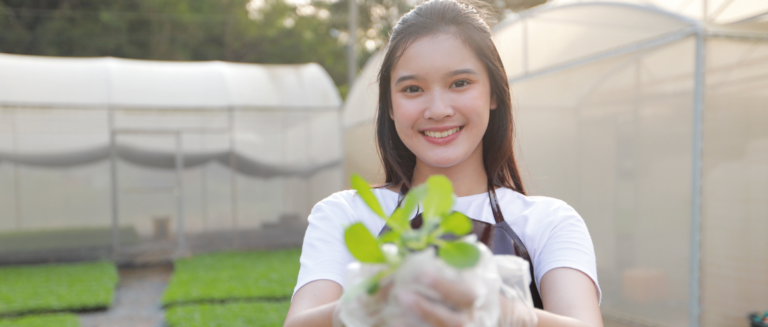 Uma mulher está sorrindo e segurando a muda de uma planta que está crescendo. Ela veste uma camiseta branca e um avental marrom. Ao fundo, tem duas estufas, representando o cultivo de alguns produtos do agronegócio no Brasil.