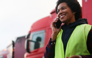 Em primeiro plano, ao lado direito, uma mulher está num pátio de veículos negociando um frete por telefone. Ao fundo tem vários caminhões estacionados.