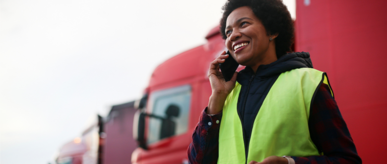 Em primeiro plano, ao lado direito, uma mulher está num pátio de veículos negociando um frete por telefone. Ao fundo tem vários caminhões estacionados.