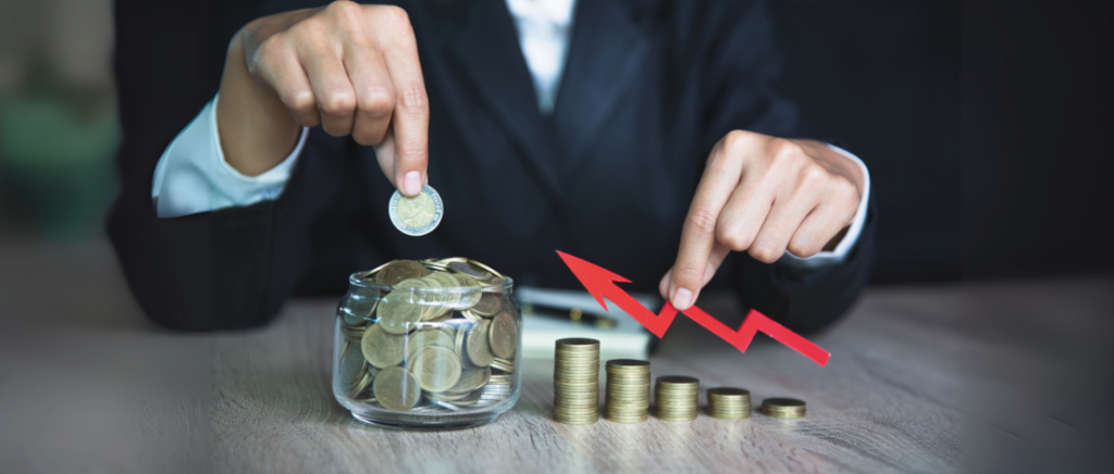 Uma pessoa está sentada e na mesa tem diversas moedas, representando a organização financeira de custos e despesas de uma empresa. Algumas moedas estão em um pote transparente e as que estão fora dele estão empilhadas. As pilhas de moedas estão em ordem decrescente. A pessoa segura uma moeda em uma mão e uma seta em ascensão na outra mão. Essa seta está sobre as moedas empilhadas, começando da direita para a esquerda, representando o aumento de ganhos.