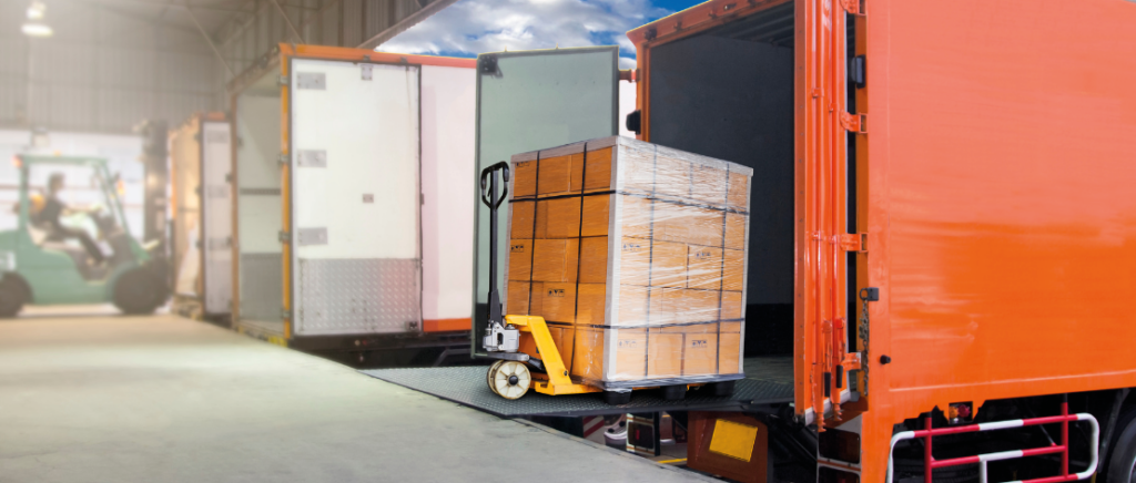 Vários caminhões estão sendo carregados com produtos no pátio de uma transportadora. No primeiro plano, um caminhão laranja está sendo carregado com uma carga de várias caixas amarelas. A imagem representa quais foram os produtos mais transportados em 2024.