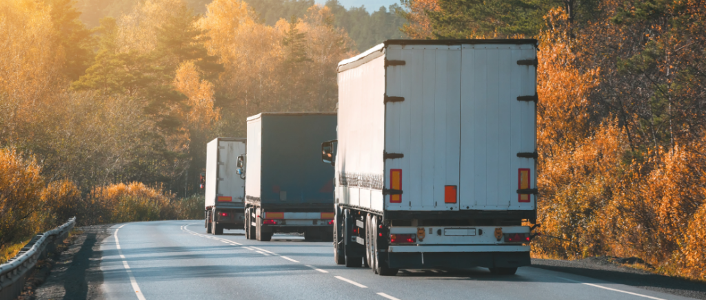 Três caminhões de carrocerias fechadas estão transportando produtos em uma estrada de faixa única. O local é repleto de árvores dos dois lados e não há outros veículos na pista.
