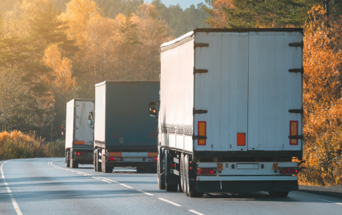 Três caminhões de carrocerias fechadas estão transportando produtos em uma estrada de faixa única. O local é repleto de árvores dos dois lados e não há outros veículos na pista.