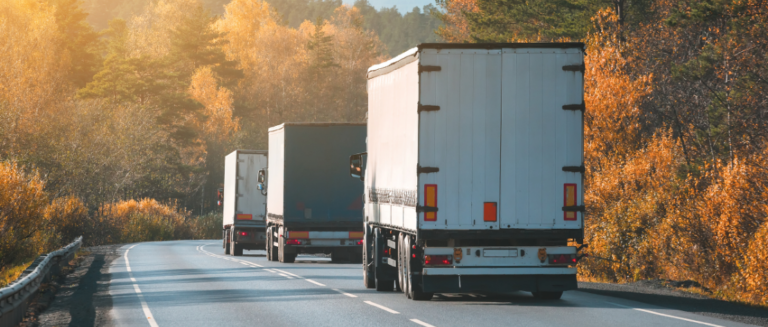 Três caminhões de carrocerias fechadas estão transportando produtos em uma estrada de faixa única. O local é repleto de árvores dos dois lados e não há outros veículos na pista.