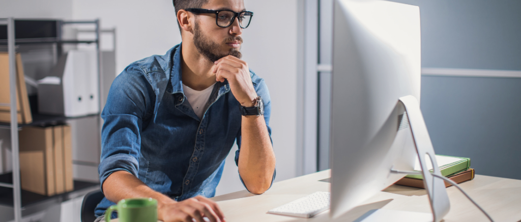 Um homem está mexendo em um computador, representando a validação e confirmação dos dados do motorista pela Fretebras. Ele está sentado e, sobre a mesa, tem um algumas pastas, o computador, o mouse e uma caneca. Ele está vestindo uma camiseta branca e uma camisa jeans. Além disso, ele tem barba e está usando óculos e um relógio analógico, ambos de cor preta.