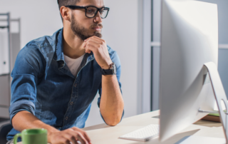 Um homem está mexendo em um computador, representando a validação e confirmação dos dados do motorista pela Fretebras. Ele está sentado e, sobre a mesa, tem um algumas pastas, o computador, o mouse e uma caneca. Ele está vestindo uma camiseta branca e uma camisa jeans. Além disso, ele tem barba e está usando óculos e um relógio analógico, ambos de cor preta.