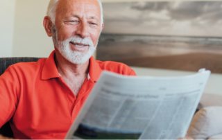 Um homem lê o jornal em sua sala, representando uma pessoa vendo as novidades de 2025 para os caminhoneiros e as transportadoras. É um senhor grisalho, com cabelo e barba bem aparados. Seu semblante é tranquilo. Ele está vestindo uma camisa polo de cor laranja e está sentado em uma poltrona.