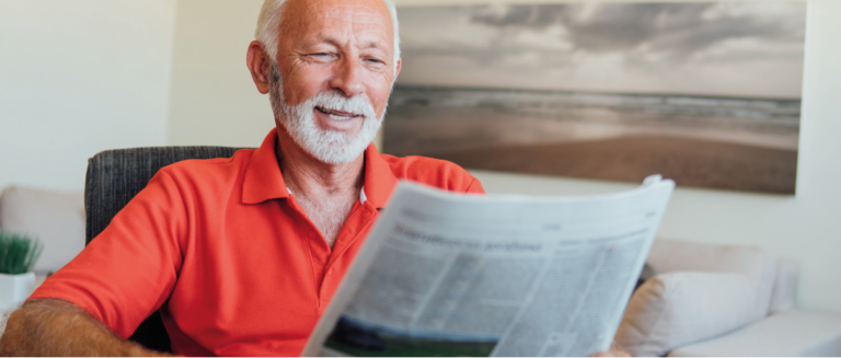 Um homem lê o jornal em sua sala, representando uma pessoa vendo as novidades de 2025 para os caminhoneiros e as transportadoras. É um senhor grisalho, com cabelo e barba bem aparados. Seu semblante é tranquilo. Ele está vestindo uma camisa polo de cor laranja e está sentado em uma poltrona.