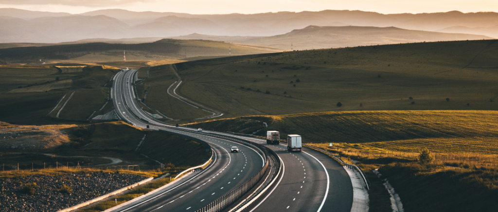 Uma estrada com vários caminhões e carros circulando, representando o transporte rodoviário. Ela tem duas faixas em cada sentido e um acostamento de cada lado. Tanto ao lado esquerdo como o direito da pista tem um gramado. No horizonte, ao fundo, tem montanhas.