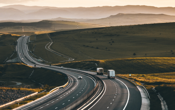 Uma estrada com vários caminhões e carros circulando, representando o transporte rodoviário. Ela tem duas faixas em cada sentido e um acostamento de cada lado. Tanto ao lado esquerdo como o direito da pista tem um gramado. No horizonte, ao fundo, tem montanhas.