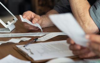 Duas pessoas estão sentadas e, sobre a mesa, tem um tablet, uma calculadora, uma caneta preta sobre uma prancheta com um papel e vários outros papéis de contas, representando os imprevistos de uma transportadora. Elas estão analisando essas informações e cada uma está segurando um papel na mão.