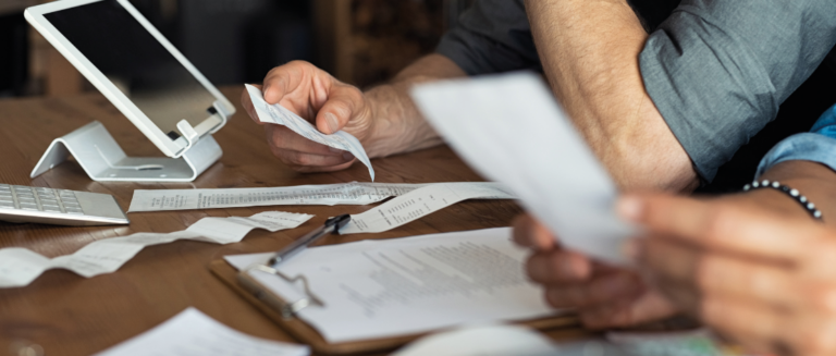 Duas pessoas estão sentadas e, sobre a mesa, tem um tablet, uma calculadora, uma caneta preta sobre uma prancheta com um papel e vários outros papéis de contas, representando os imprevistos de uma transportadora. Elas estão analisando essas informações e cada uma está segurando um papel na mão.