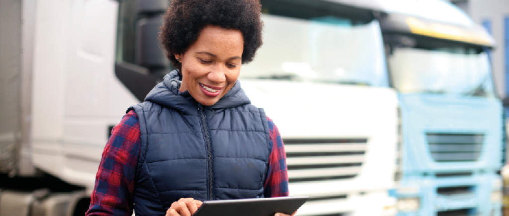 A imagem mostra uma mulher, em primeiro plano, representando o empreendedorismo feminino. Ela está ao lado esquerdo, com um tablet nas mãos, e está resolvendo assuntos da sua transportadora. Em segundo plano, desfocado, tem vários caminhões que representam sua frota.