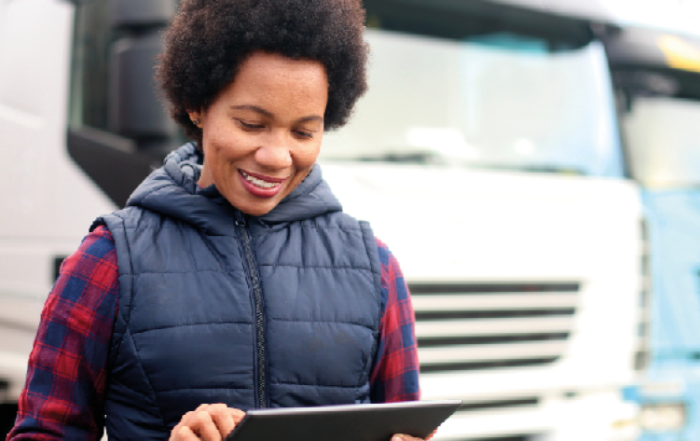 A imagem mostra uma mulher, em primeiro plano, representando o empreendedorismo feminino. Ela está ao lado esquerdo, com um tablet nas mãos, e está resolvendo assuntos da sua transportadora. Em segundo plano, desfocado, tem vários caminhões que representam sua frota.