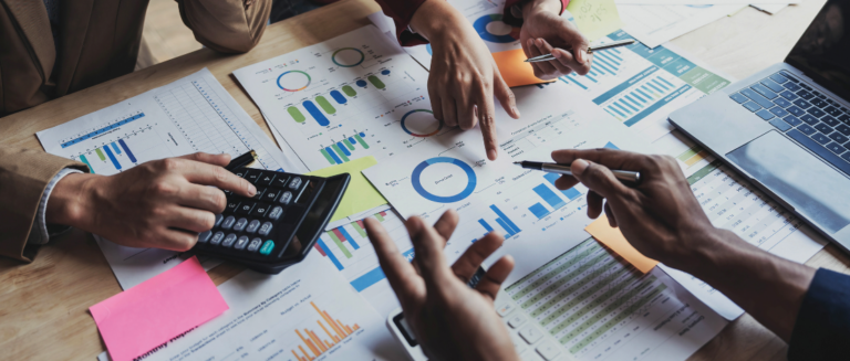 Três pessoas estão sentadas e analisando vários dados nos papéis que estão sobre a mesa, representando a gestão financeira de uma transportadora. Duas pessoas estão segurando canetas e apontando para os gráficos, enquanto a outra faz contas na calculadora.