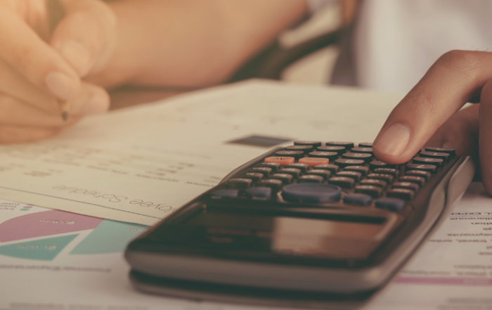 Uma pessoa está fazendo contas em uma mesa, representando um caminhoneiro calculando os custos das cargas. Ele usa uma calculadora e segura um lápis. Também tem vários papéis sobre a mesa e o motorista está fazendo anotações neles.