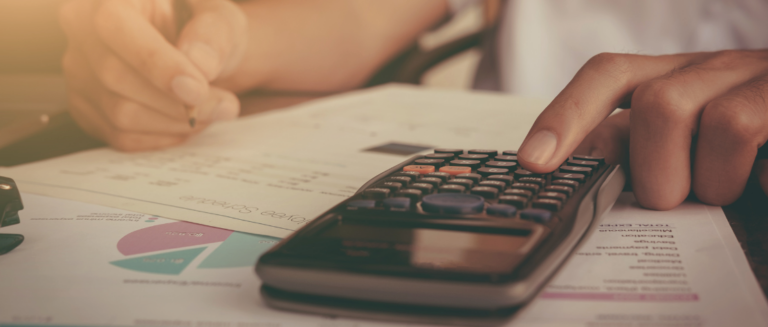 Uma pessoa está fazendo contas em uma mesa, representando um caminhoneiro calculando os custos das cargas. Ele usa uma calculadora e segura um lápis. Também tem vários papéis sobre a mesa e o motorista está fazendo anotações neles.
