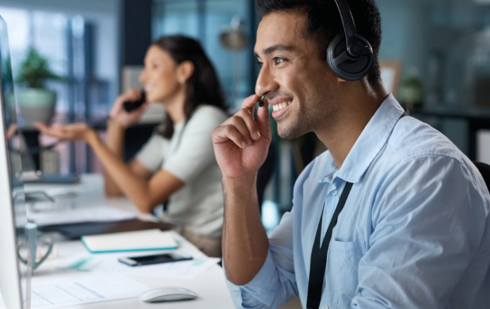 Um homem e uma mulher estão atendendo clientes por ligação. O homem está em primeiro plano, usando um headphone. Já a mulher, em segundo plano, está utilizando um telefone. Ambos estão sorridentes e com computadores e papéis de apoio.