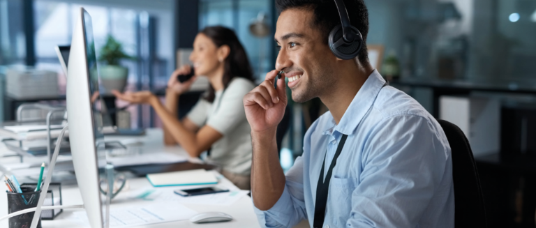 Um homem e uma mulher estão atendendo clientes por ligação. O homem está em primeiro plano, usando um headphone. Já a mulher, em segundo plano, está utilizando um telefone. Ambos estão sorridentes e com computadores e papéis de apoio.