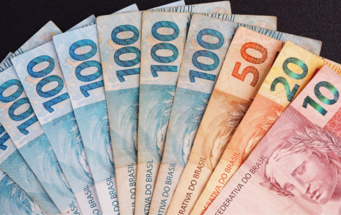 A imagem mostra várias notas sobre um fundo preto, representando o crédito para as transportadoras. Tem uma nota de R$10, uma de R$20, uma de R$50 e oito de R$100, totalizando a quantia de R$880.