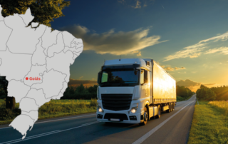 A imagem mostra um caminhão em uma estrada. Os dois lados da rodovia têm grama e algumas árvores. Em primeiro plano, ao lado esquerdo, tem um mapa do Brasil, com identificação do estado de Goiás, representando os caminhões usados em fretes para Goiás.