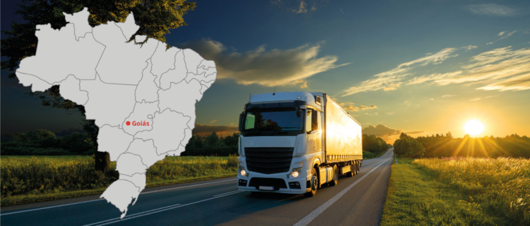 A imagem mostra um caminhão em uma estrada. Os dois lados da rodovia têm grama e algumas árvores. Em primeiro plano, ao lado esquerdo, tem um mapa do Brasil, com identificação do estado de Goiás, representando os caminhões usados em fretes para Goiás.