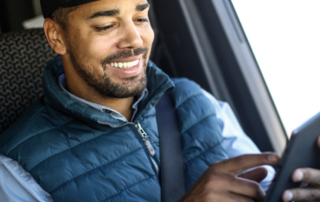 A imagem mostra um motorista dentro de seu caminhão. Ele está sentado no banco, usando o cinto de segurança e o veículo está parado. O motorista está mexendo em um tablet, representando a avaliação da empresa dentro da plataforma da Fretebras.