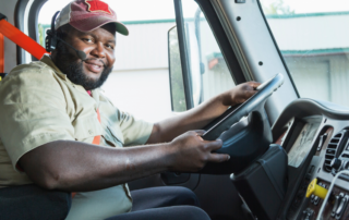 A imagem mostra um motorista dentro do seu caminhão. Ele está sentado, com cinto de segurança e com as mãos sobre o volante. Seu semblante está feliz, representando os motoristas que usam e aprovam a Fretebras.