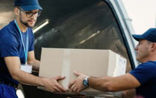 Dois entregadores vestindo uniformes azuis movimentando uma caixa de papelão, mostrando a logística de transporte de mercadorias nas modalidades de frete CIF e FOB.