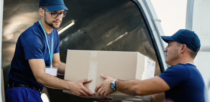 Dois entregadores vestindo uniformes azuis movimentando uma caixa de papelão, mostrando a logística de transporte de mercadorias nas modalidades de frete CIF e FOB.