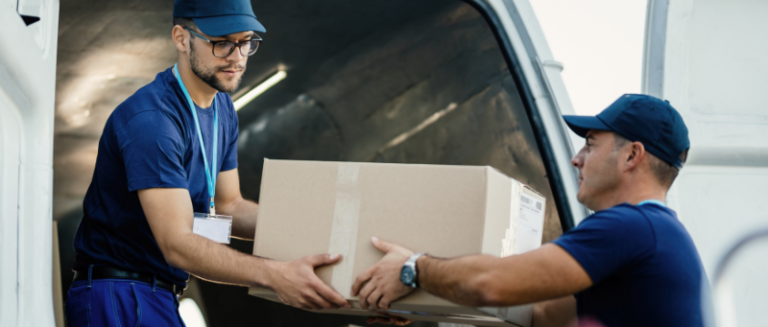 Dois entregadores vestindo uniformes azuis movimentando uma caixa de papelão, mostrando a logística de transporte de mercadorias nas modalidades de frete CIF e FOB.