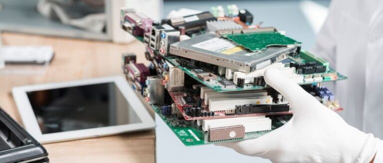 Um técnico com luvas brancas segurando uma pilha de peças eletrônicas sobre uma bancada de trabalho, ilustrando a etapa de triagem, reparo e recuperação de materiais na logística reversa de equipamentos eletrônicos.