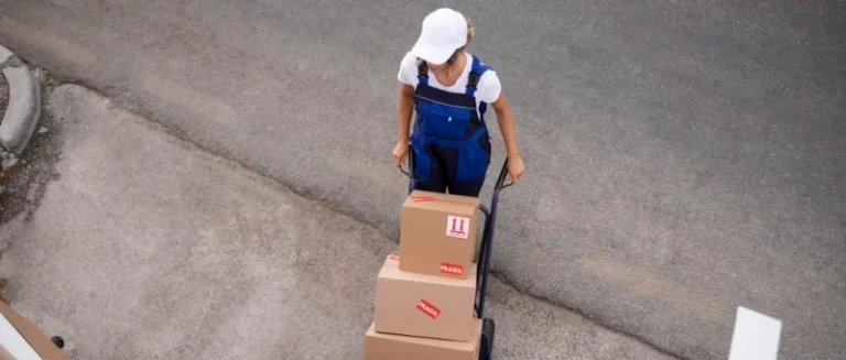 Uma entregadora uniformizada usando um carrinho de carga para transportar caixas com em direção às portas abertas de um veículo, demonstrando a etapa de coleta ou entrega em uma operação de carga fracionada